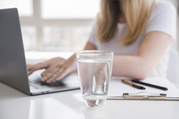 Na pierwszym planie szklanka z wodą. W tle kobieta pracująca przy laptopie.