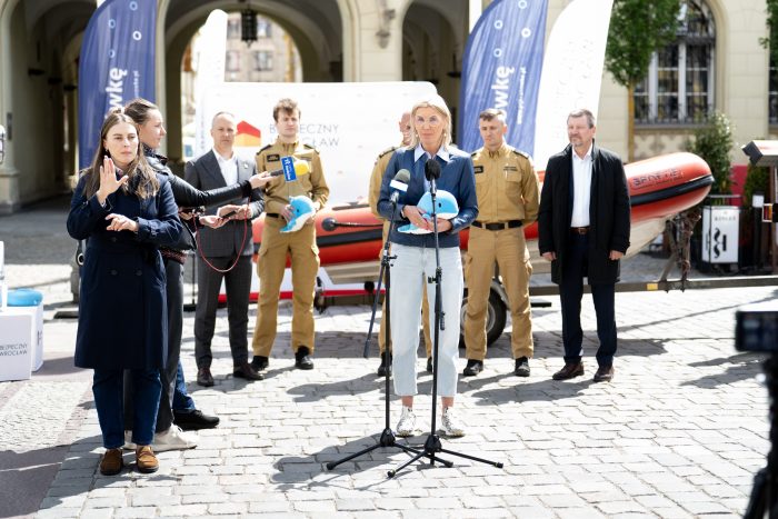 Szeroki plan konferencji przed budynkiem – osoba przemawia do mikrofonów, obok tłumaczka PJM; w tle strażacy, baner „Bezpieczny Wrocław” i ponton.