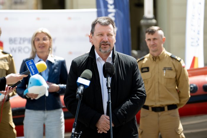 Osoba w ciemnej kurtce przemawia do mikrofonów podczas konferencji; za nią strażacy w mundurach i sprzęt ratowniczy.